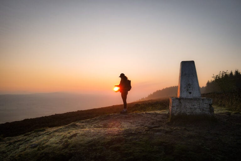 Beck stands next to the trig point during the Longridge Fell Walk at sunrise