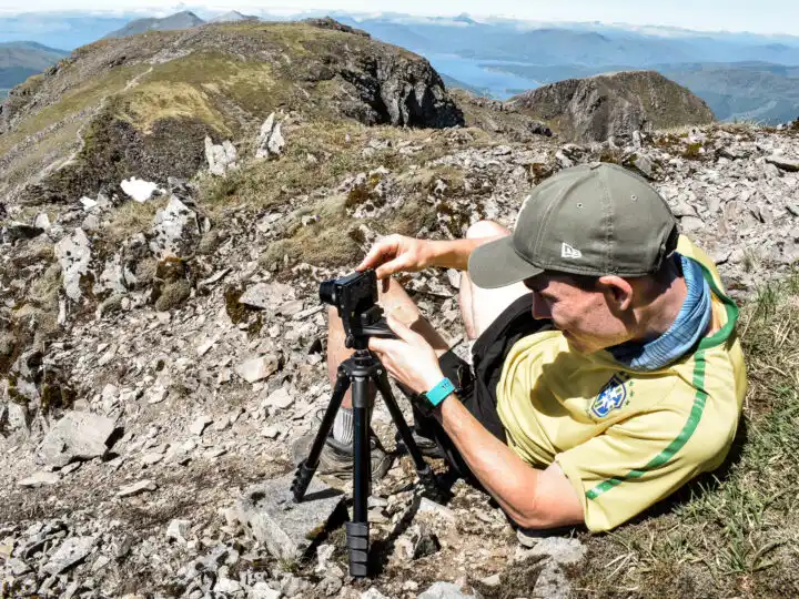 Dan is lying down, but propped up, using camera equipment to take a photo in a mountainous area