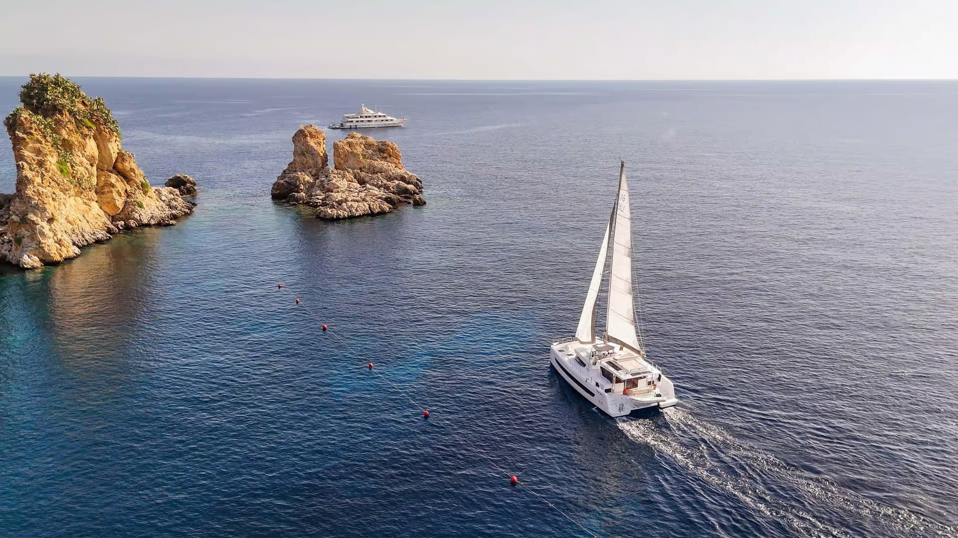 A catamaran sails towards sea stacks called Faraglioni di Scopello