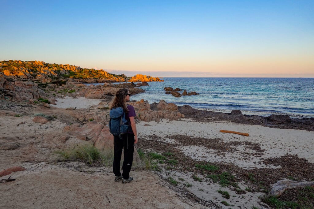 Beck stands on a rocky surface, next to a small beach on Sentier Littoral des Bruzzi