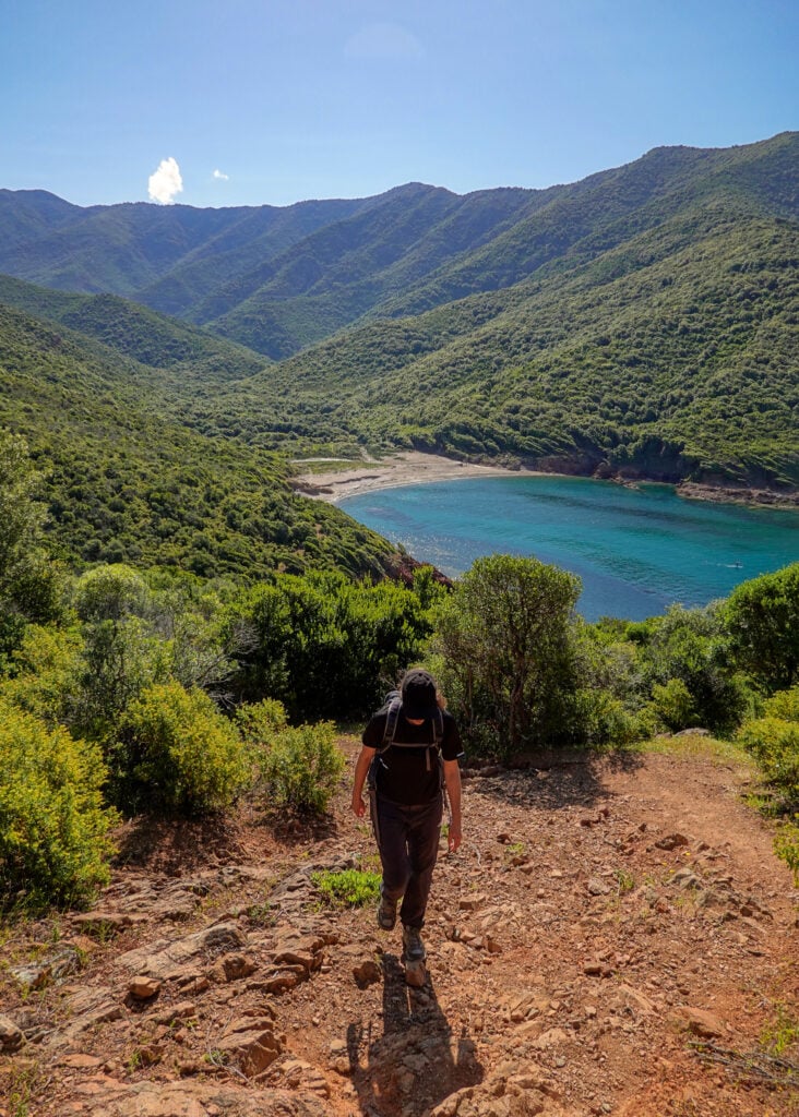 Beck hikes up a rocky trail heading away from a beach on a hike in Corsica called Sentier du Facteur Girolata