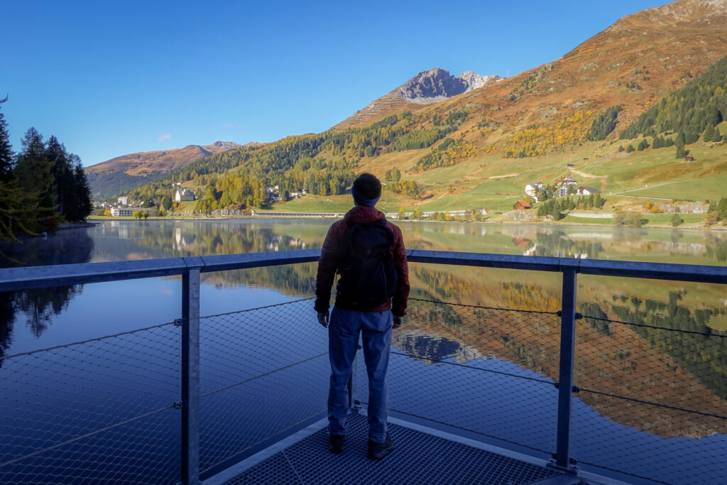 Dan stands on a viewing platform overseeing a lake surrounded by mountains during a walk called Davosersee Rundweg (AKA Lake Davos Trail)
