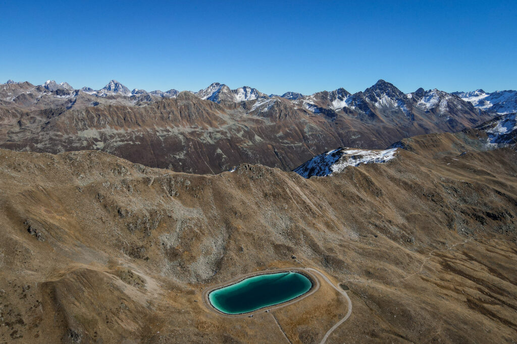 An aerial view of a small lake dwarfed by mountains taken during the Jakobshorn Hike to Sertig