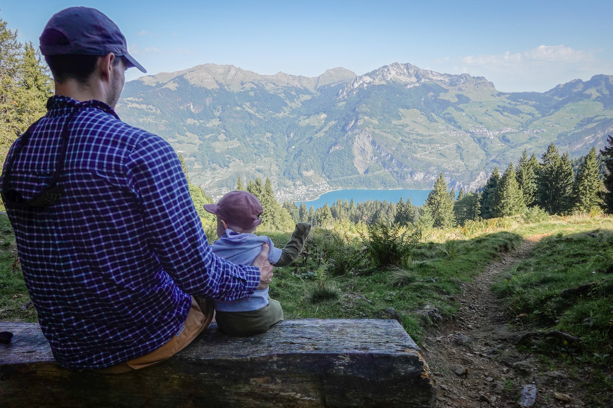 Dan and baby sit on a bench overlooking a lake and mountains during the Schabziger Höhenweg