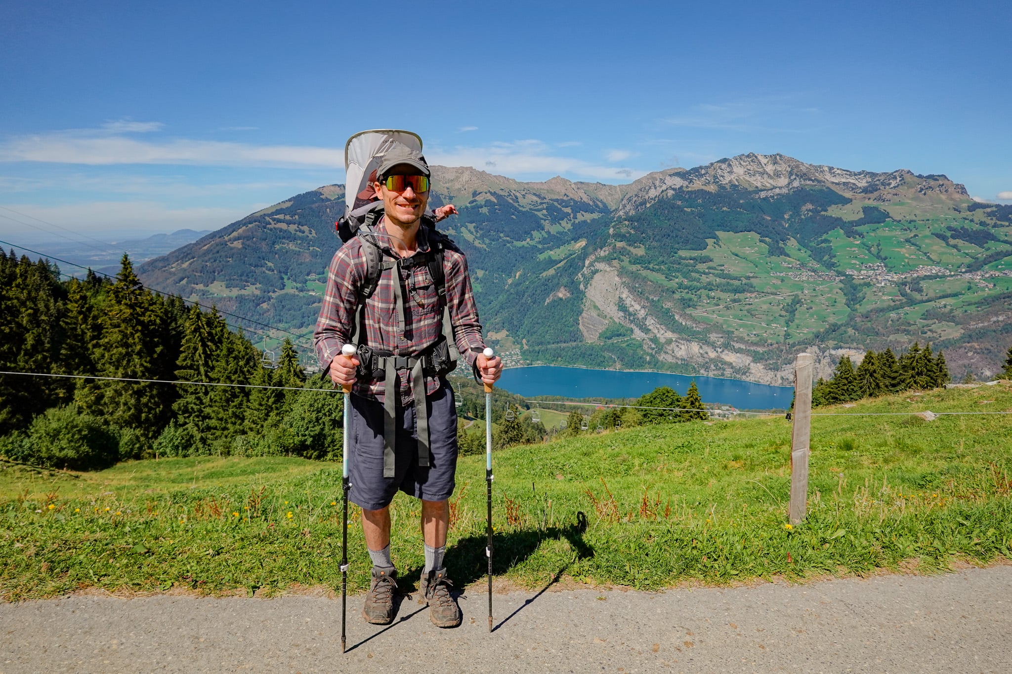 Mountains and a lake is seen behind Dan who is wearing a child carrier backpack on the Habergschwänd Talalpsee Hike